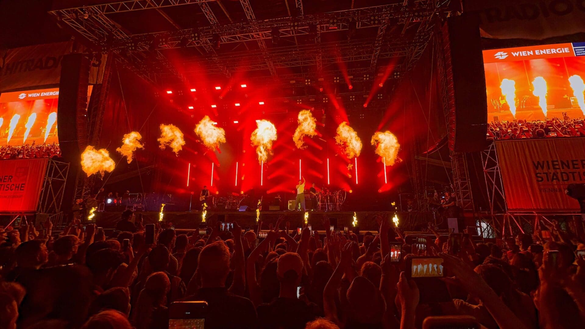 Ein Musiker steht auf einer Bühne mit Pyrotechnik und roten Scheinwerfern vor einem großen Publikum.