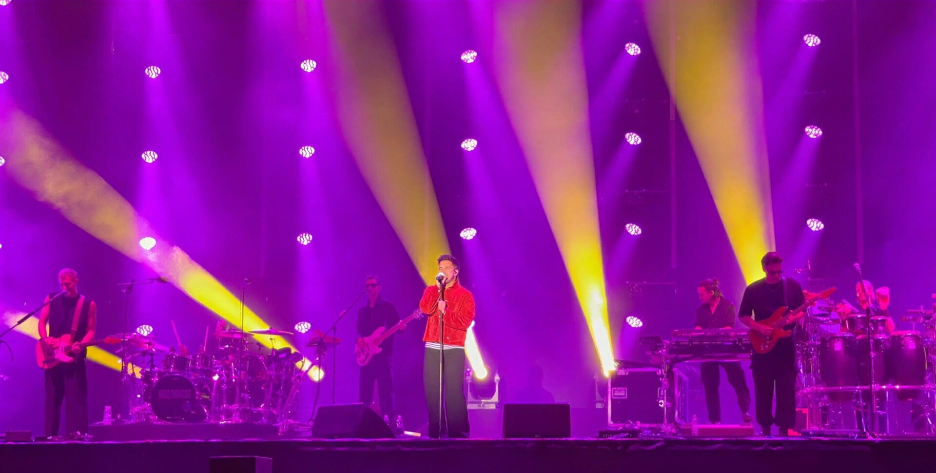 Sänger in roter Jacke singt mit Mikrofon auf lila beleuchteter Bühne vor Bandmitgliedern