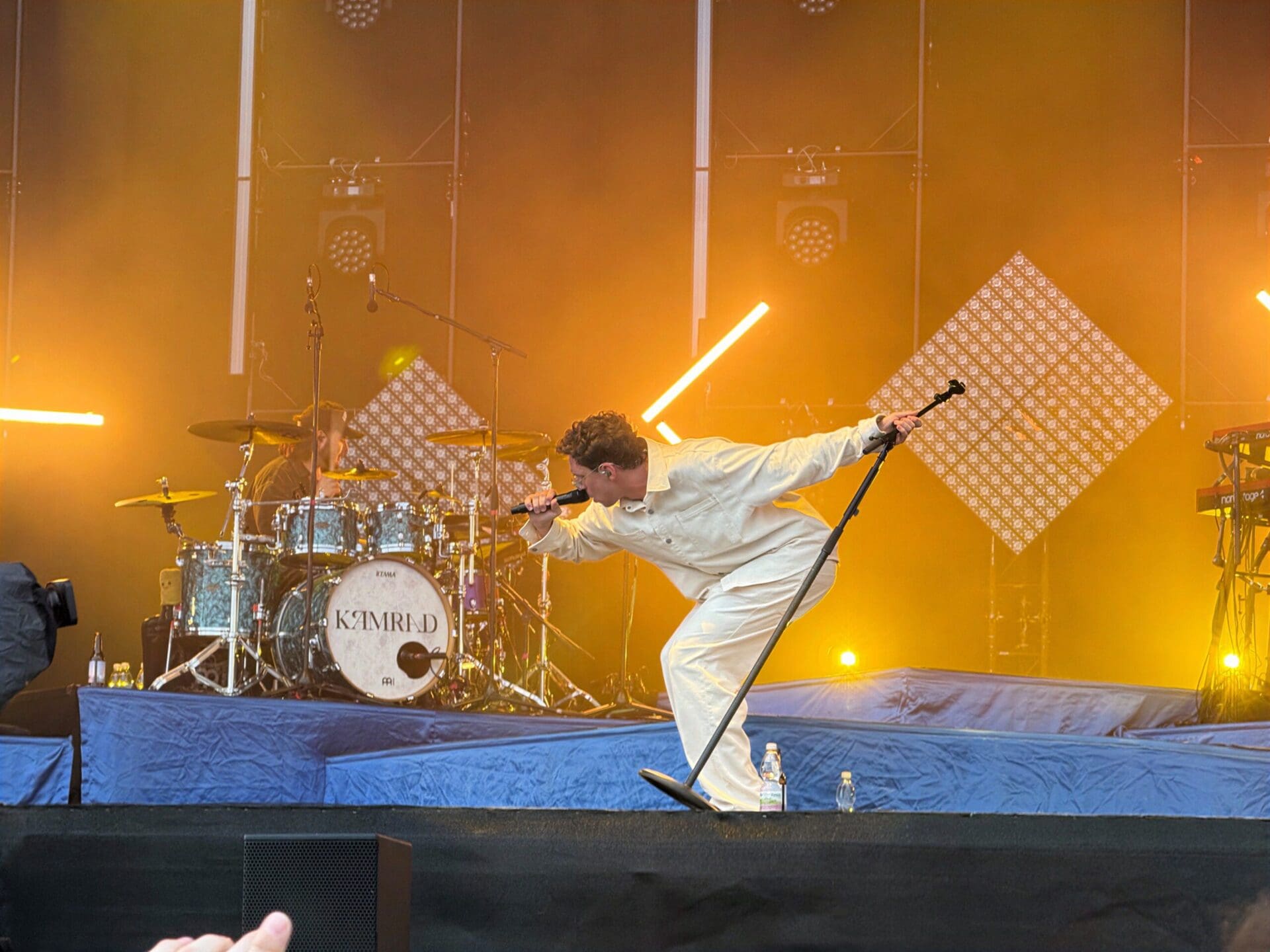 Sänger in weißem Outfit singt energisch auf der Bühne mit Mikrofon und Schlagzeug im Hintergrund beim Donauinselfest.