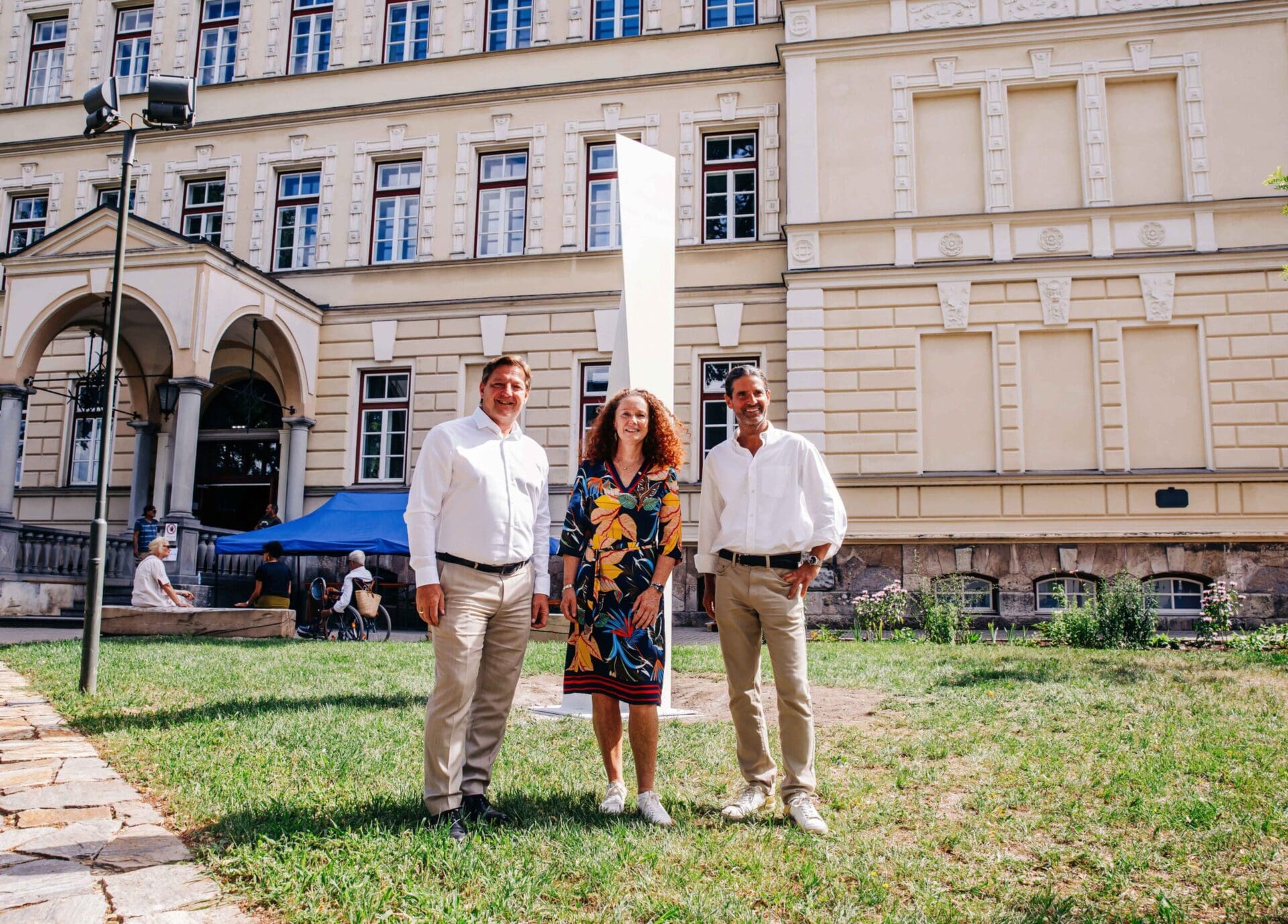 Drei Personen stehen vor einer weißen Stahlskulptur auf einer Wiese vor einem großen Gebäude.