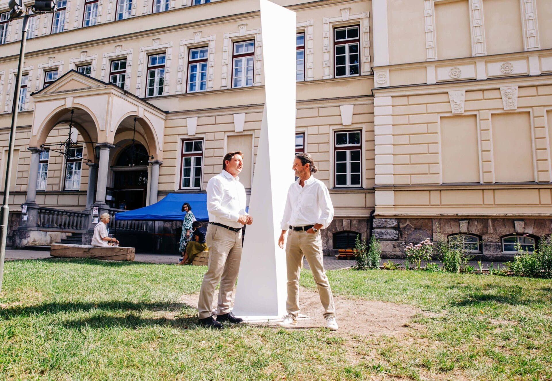 Zwei Männer stehen neben einer hohen weißen Stahlskulptur auf einer Wiese vor einem Gebäude
