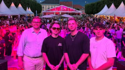 Vier Männer posieren auf einer Bühne bei einer Abendveranstaltung mit rosa Beleuchtung und Menschenmenge im Hintergrund