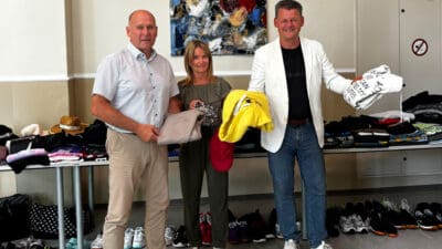 Drei Personen stehen vor Tischen mit Kleidung und Schuhen und halten Textilien in den Händen.