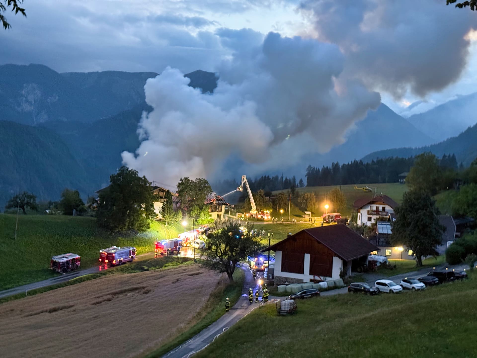 Feuerwehrleute und Einsatzfahrzeuge bekämpfen einen Vollbrand eines Wirtschaftsgebäudes mit starker Rauchwolke in den Bergen bei Dämmerung