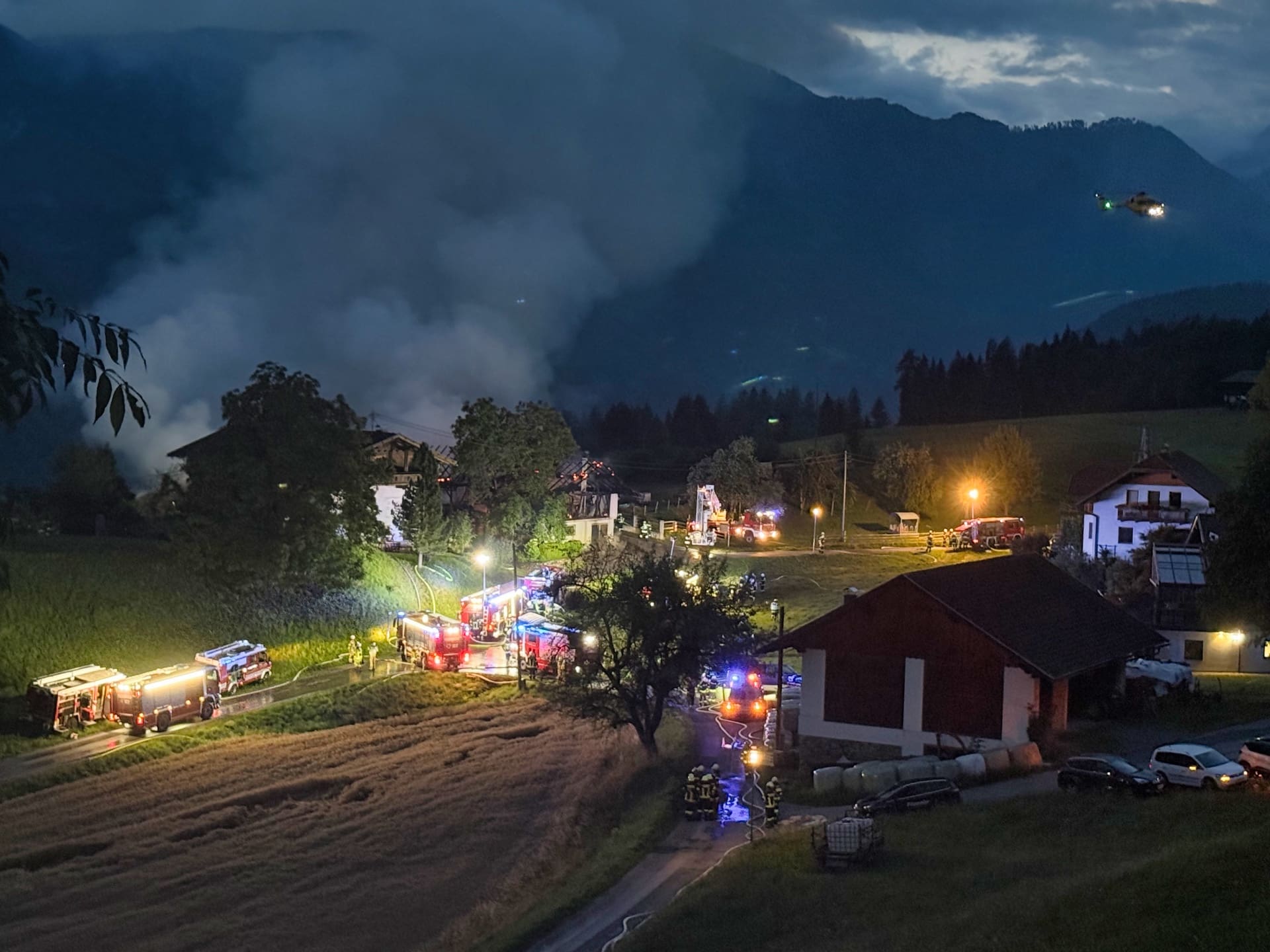 Feuerwehrfahrzeuge mit Blaulicht löschen Brand eines Wirtschaftsgebäudes bei Nacht vor Bergkulisse mit Hubschrauber.