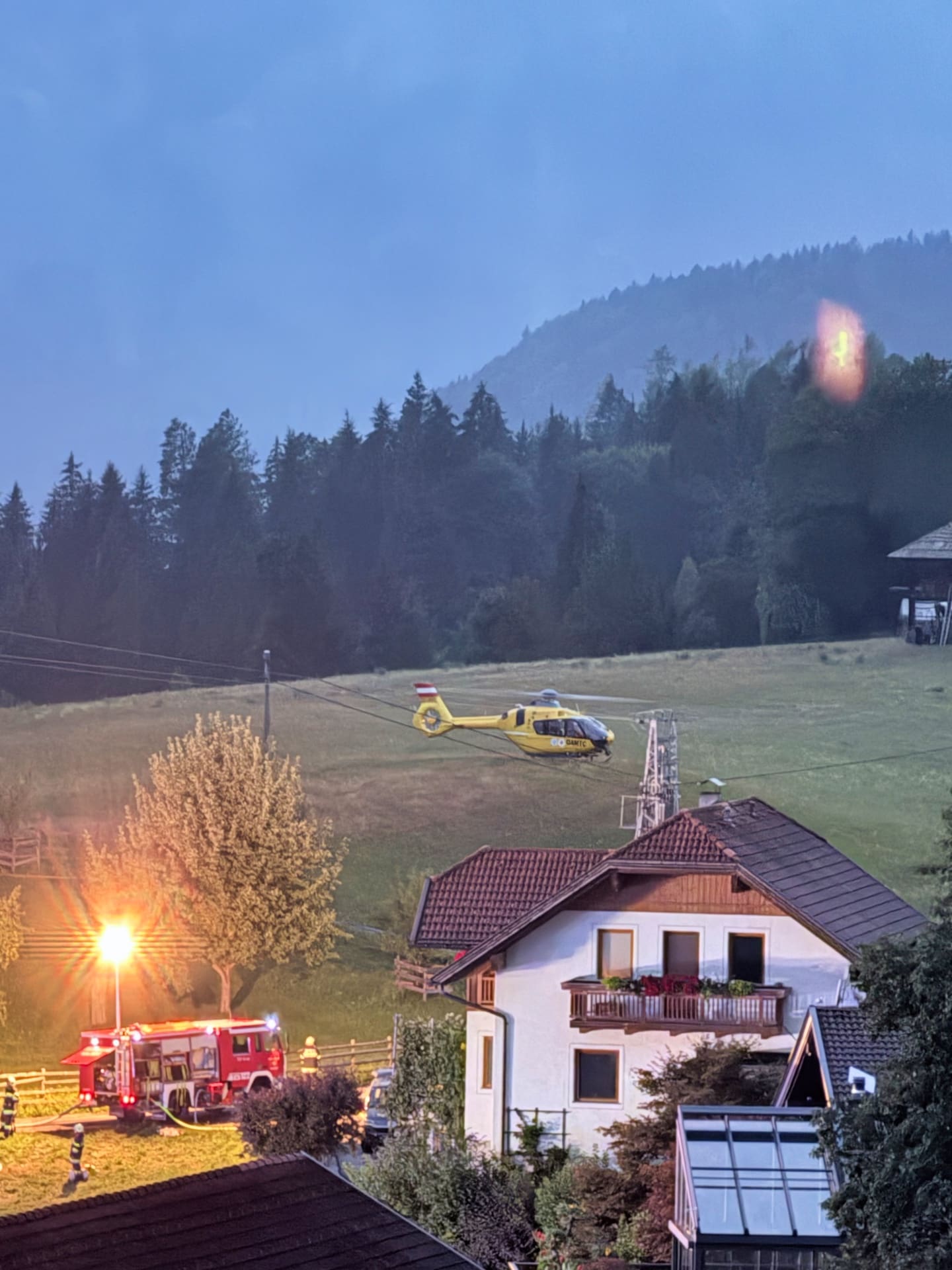 Gelber Rettungshubschrauber fliegt über einer Wiese neben rotem Feuerwehrfahrzeug und Haus