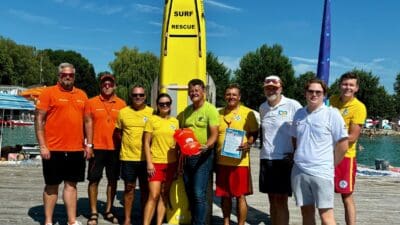 Gruppe von Rettungskräften in bunten T-Shirts steht vor einem gelben Surfrettungsbrett auf einem Holzsteg am Wasser.