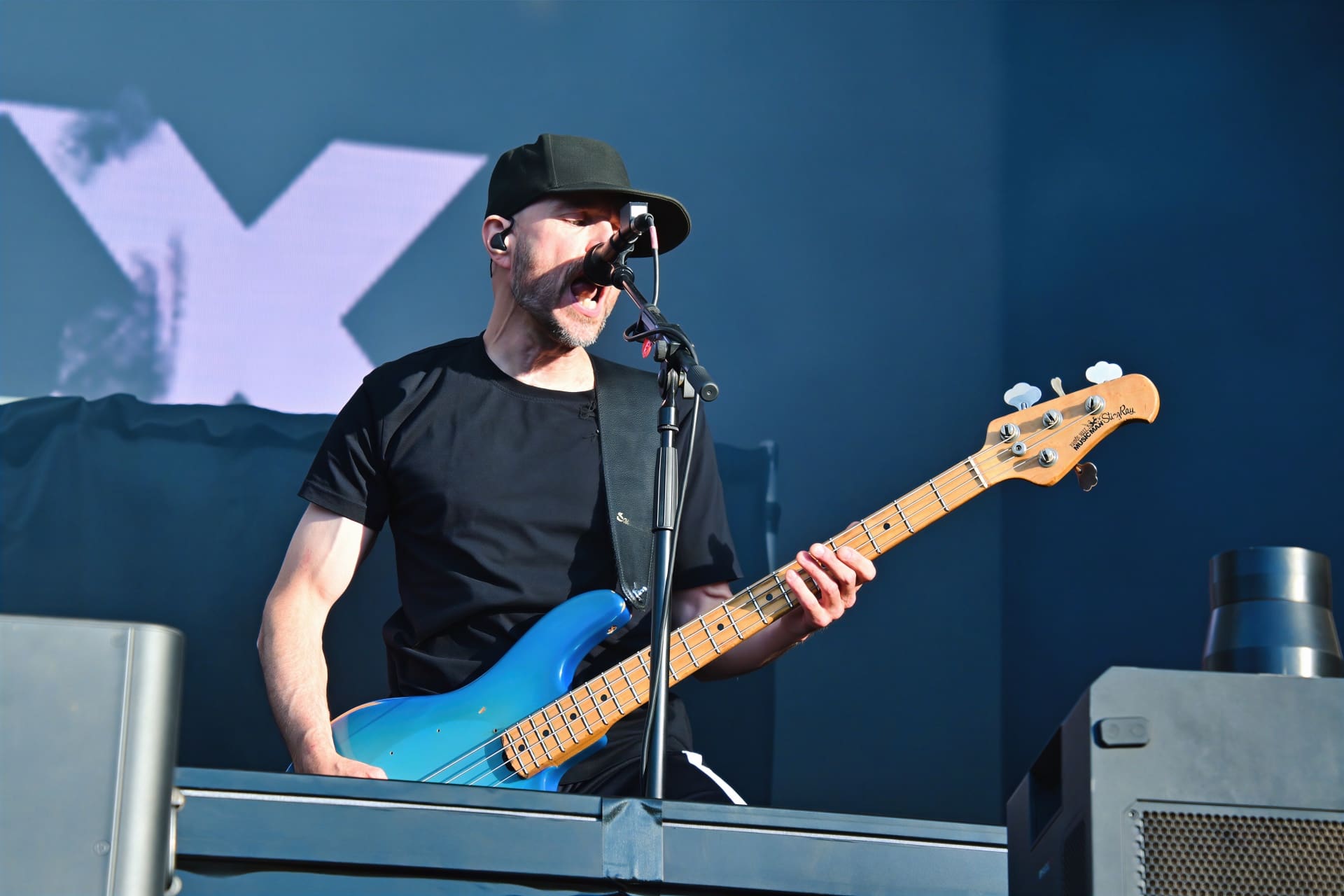 Ein Bassist mit schwarzer Kappe spielt auf einer blauen Bassgitarre und singt ins Mikrofon.