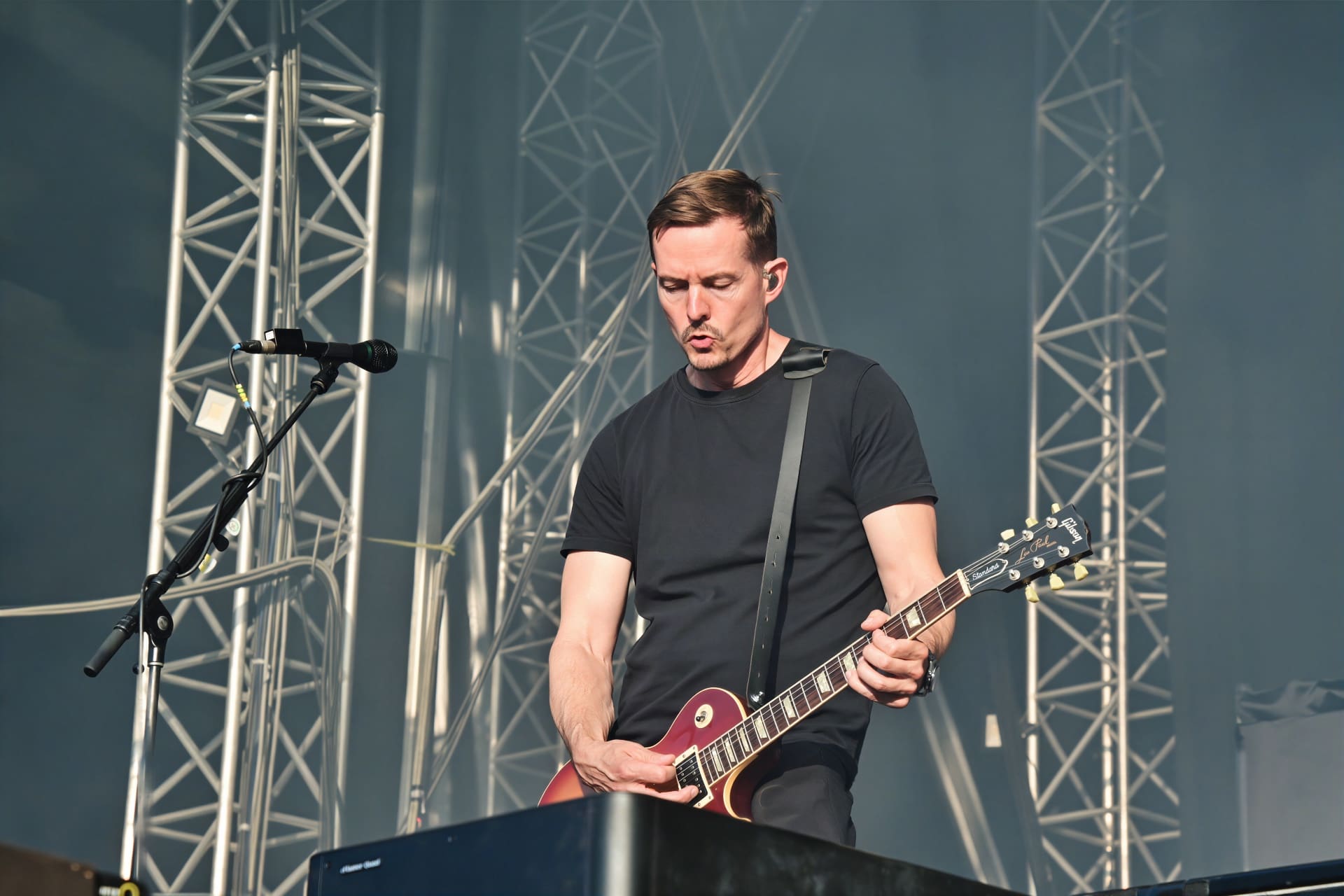 Ein Gitarrist mit kurzen braunen Haaren spielt eine rote E-Gitarre auf einer Bühne mit Metallgerüst.