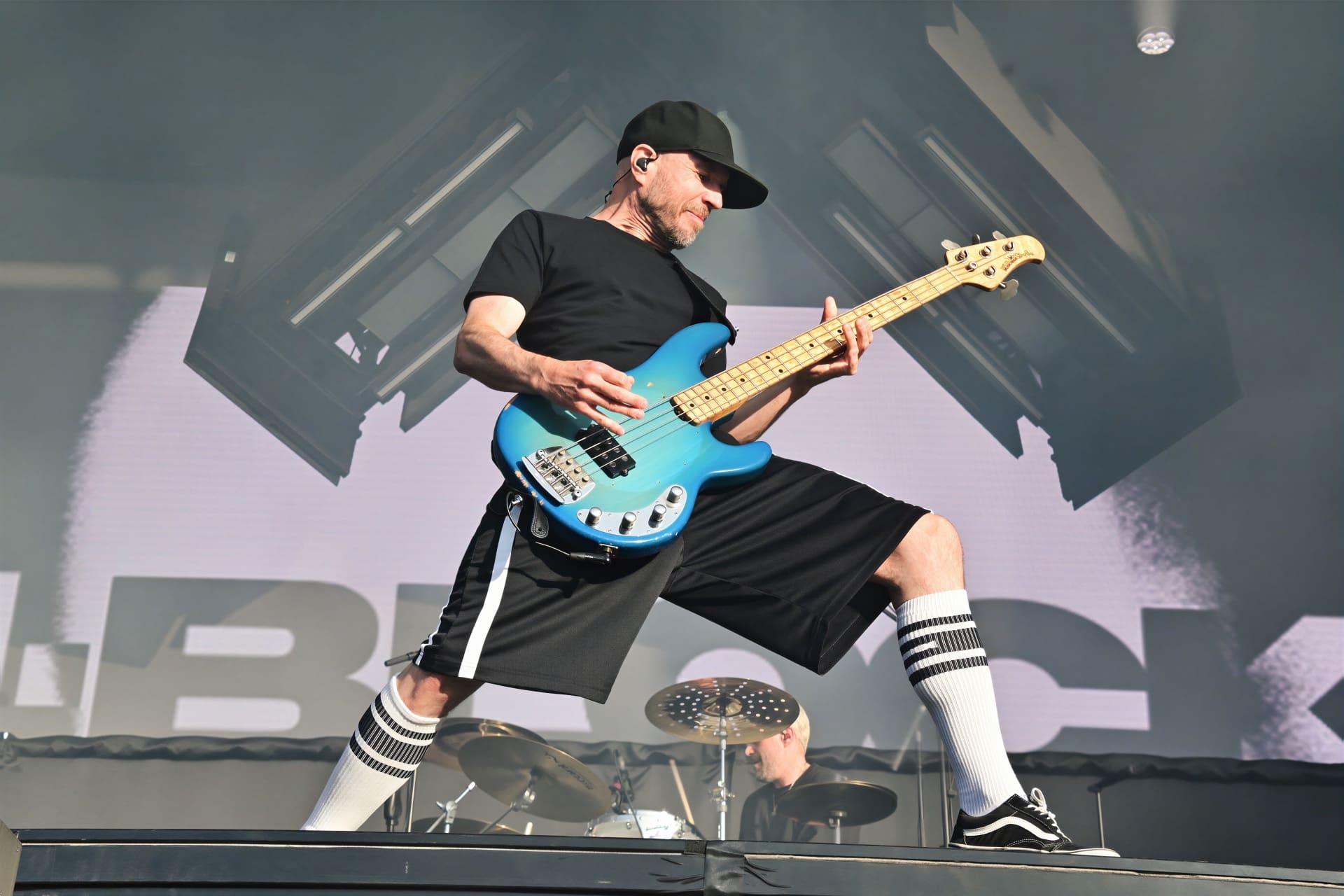 Ein Bassist mit schwarzer Kappe spielt eine blaue Bassgitarre auf einer verrauchten Konzertbühne.