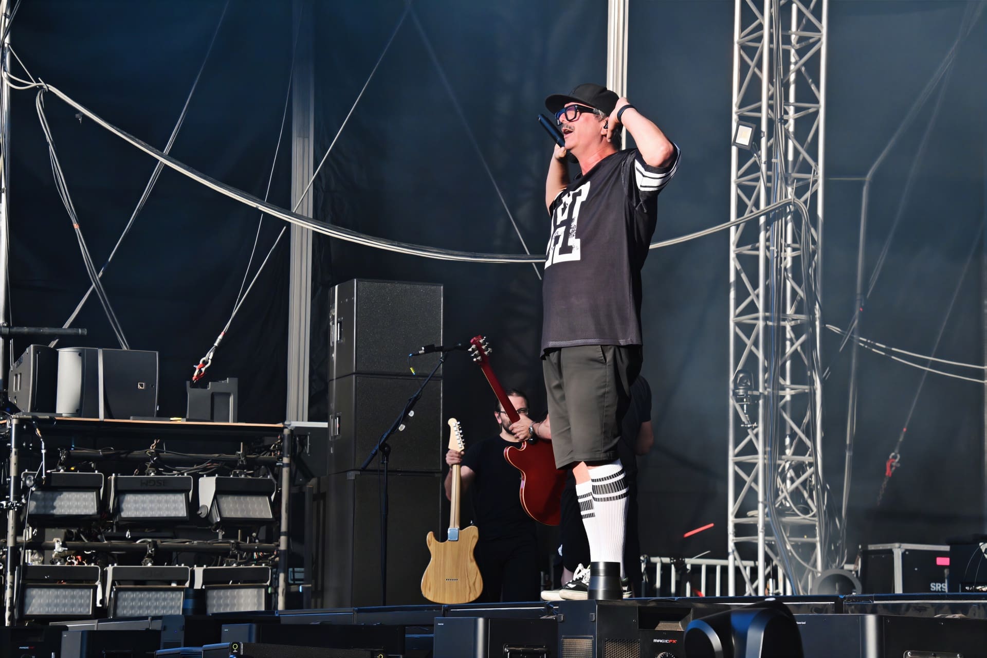 Sänger mit schwarzer Cap und Brille singt in Mikrofon auf Konzertbühne mit technischer Ausstattung