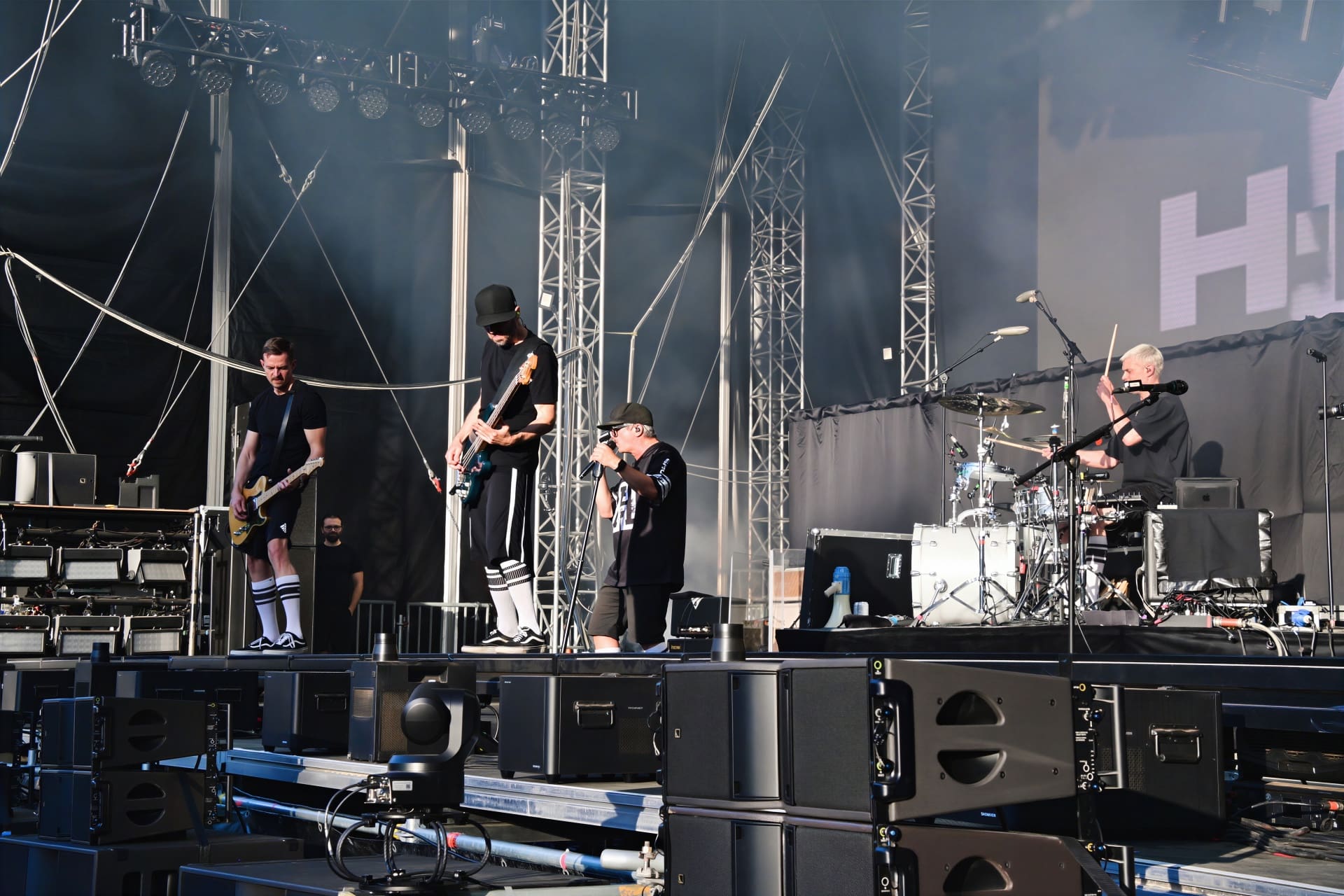 Musiker mit E-Gitarre, Bass, Mikrofon und Schlagzeug auf einer Konzertbühne mit Rauch und Lichteffekten
