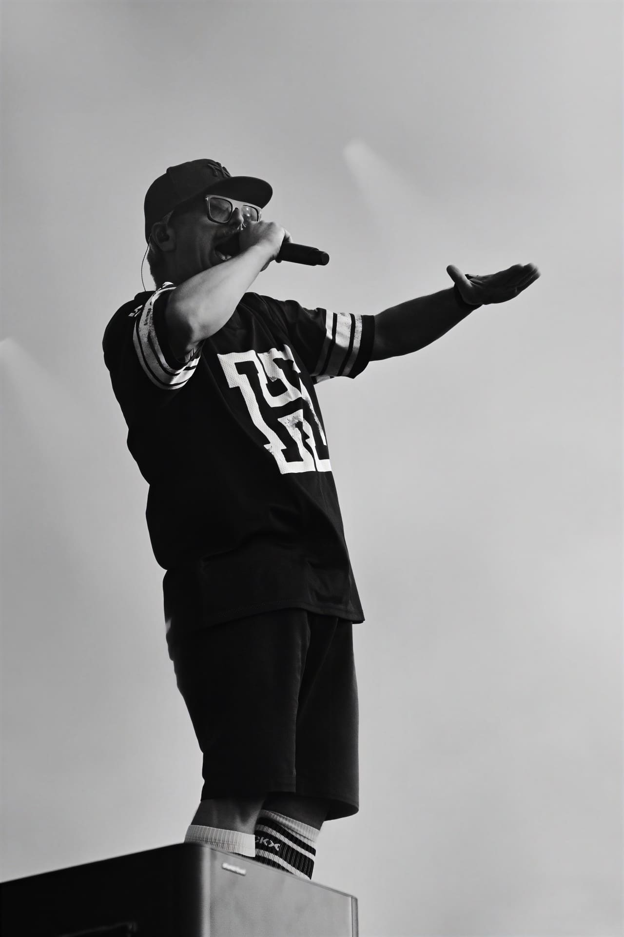 Schwarz-weißes Foto eines Sängers auf der Bühne mit Cap und Brille, der mit ausgestrecktem Arm ins Mikrofon singt.