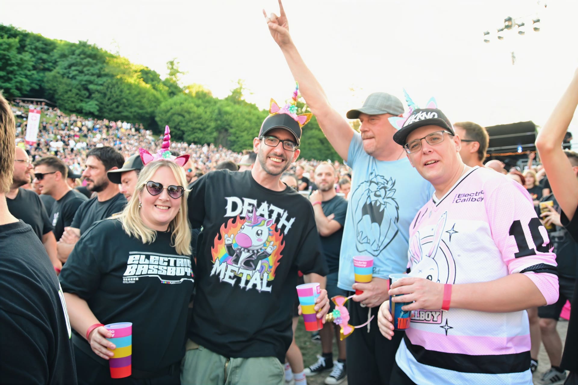 Fans mit Einhorn-Stirnbändern und Electric-Callboy-Merch tragen bunte Becher vor einer großen Menschenmenge.