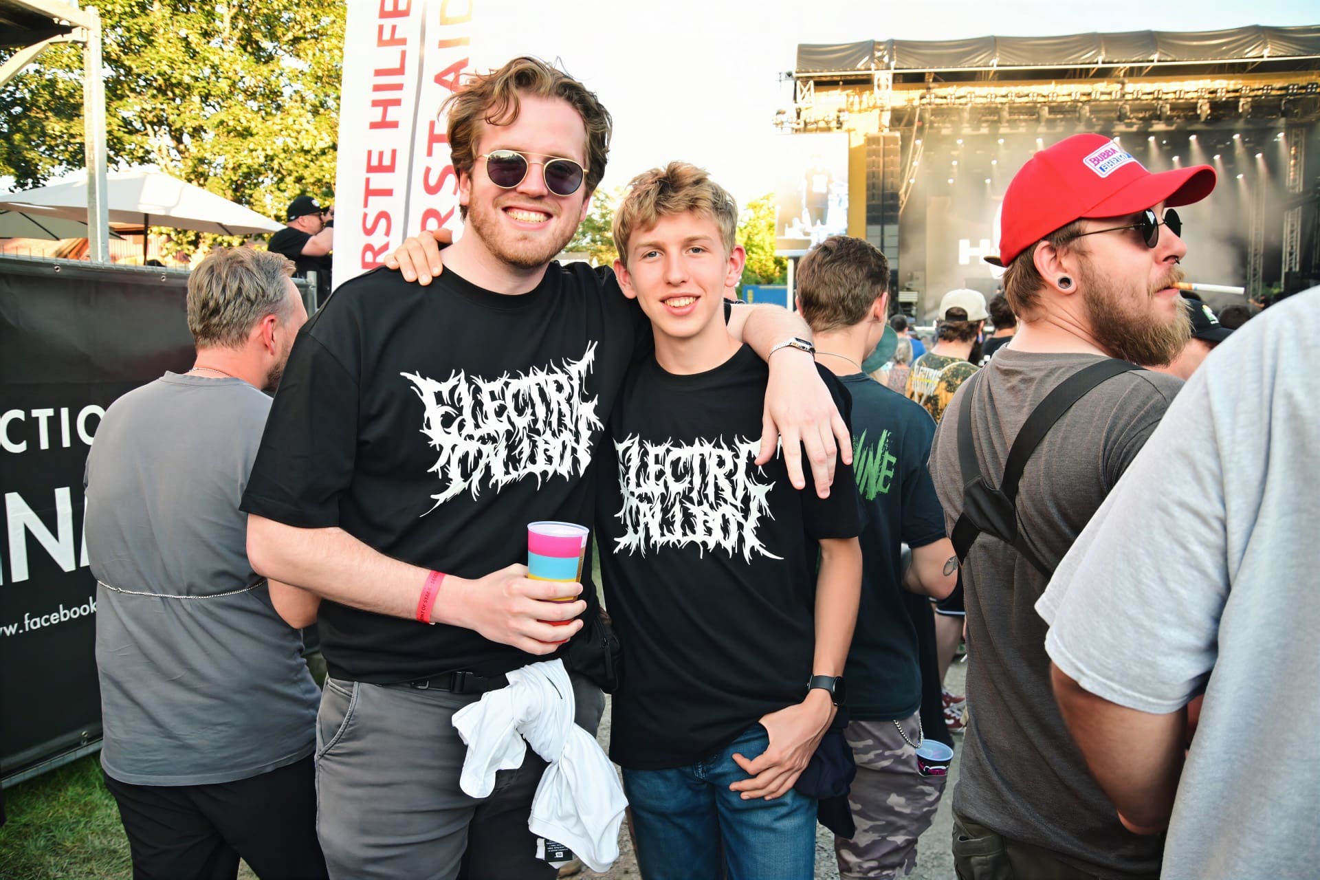 Zwei lächelnde junge Männer mit Electric Callboy T-Shirts umarmen sich vor der Bühne.