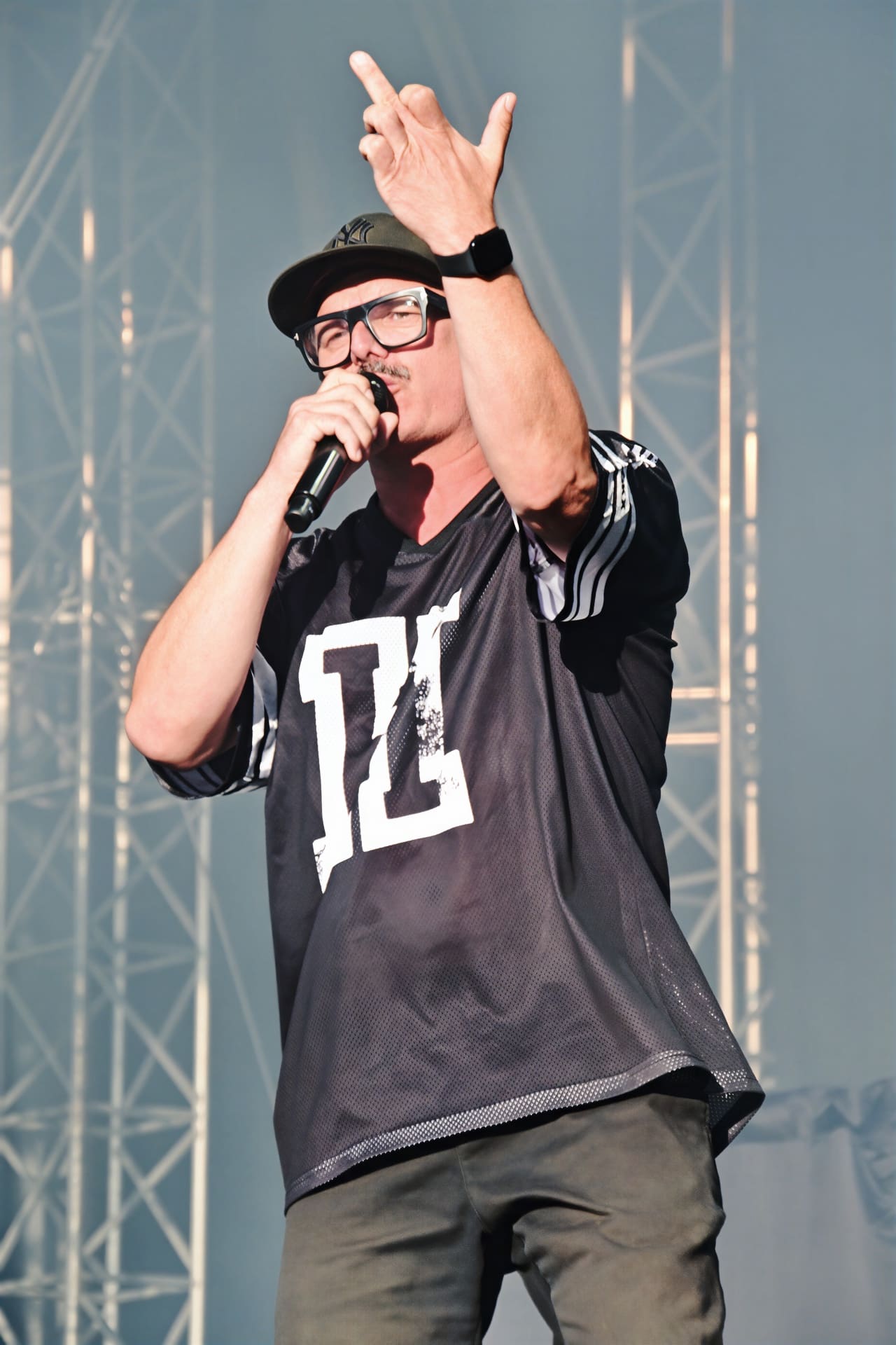 Ein Sänger mit Brille und Cap singt in ein Mikrofon und hebt die Hand auf der Bühne.