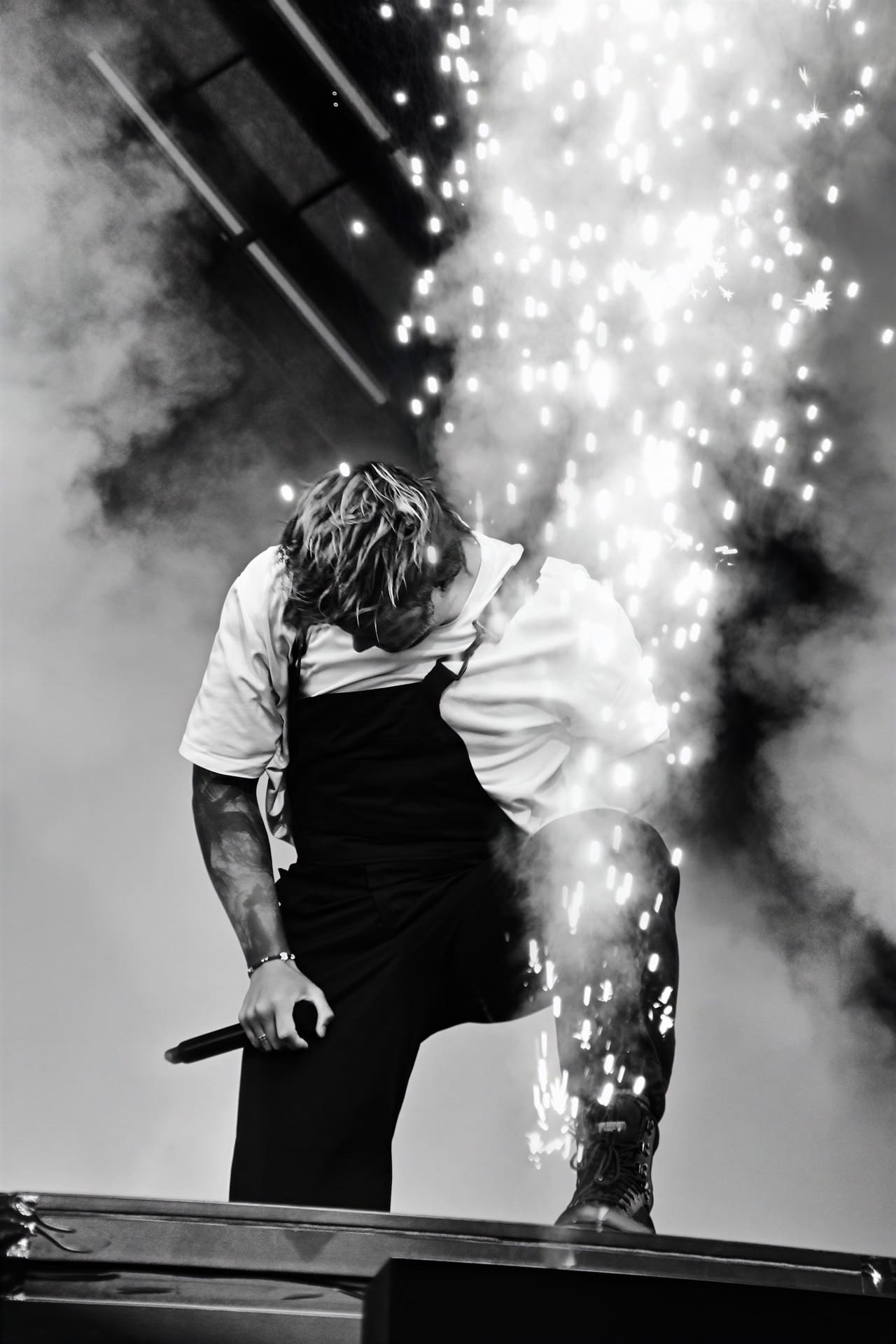 Männlicher Sänger in weißem Shirt und schwarzer Latzhose beugt sich auf Bühne mit Pyrotechnik und Funken.