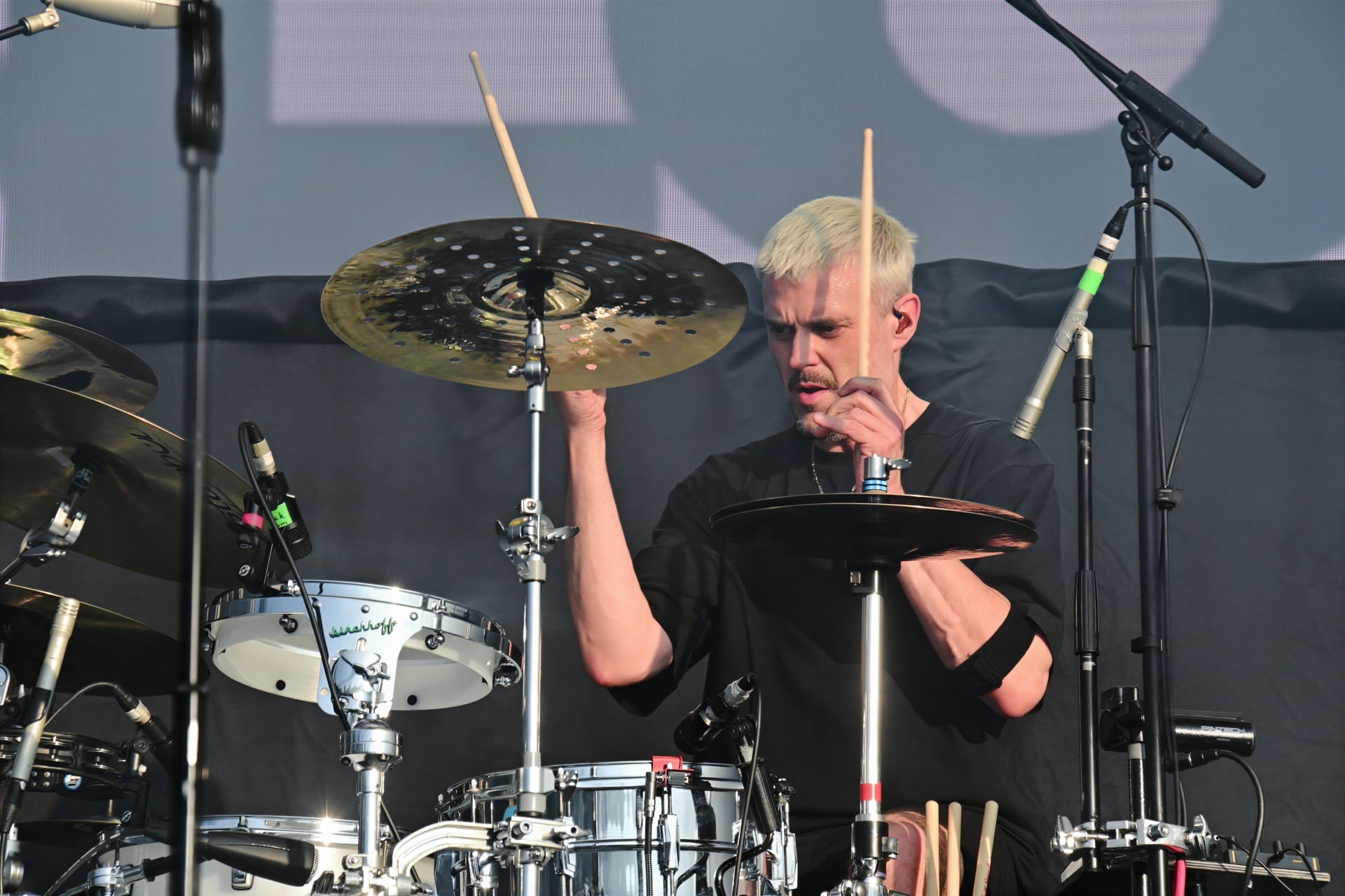 Ein Mann mit hellblondem Haar spielt Schlagzeug auf einer Konzertbühne und hält zwei Drumsticks.