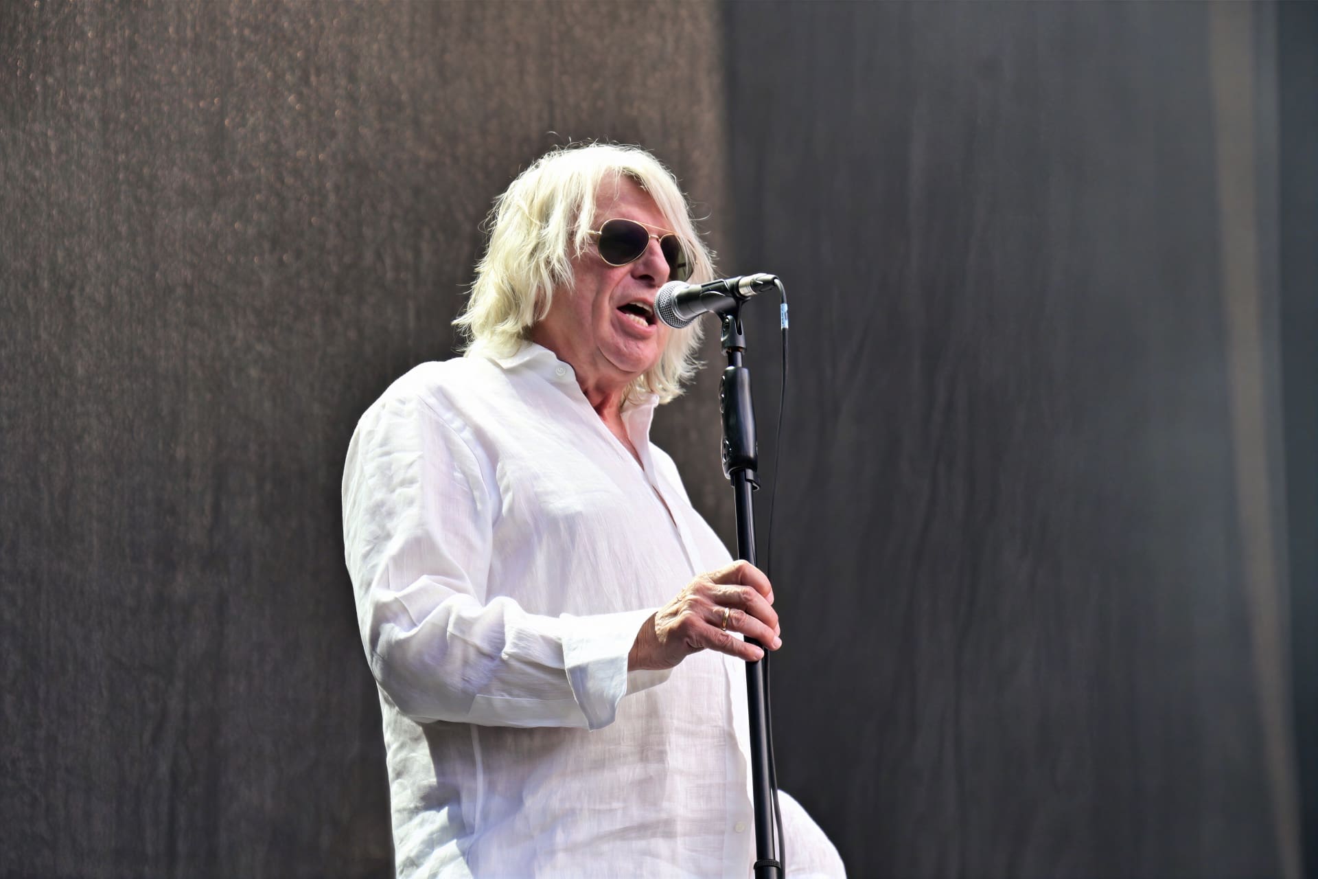 Ein älterer Sänger mit langen weißen Haaren und Sonnenbrille singt in ein Mikrofon.