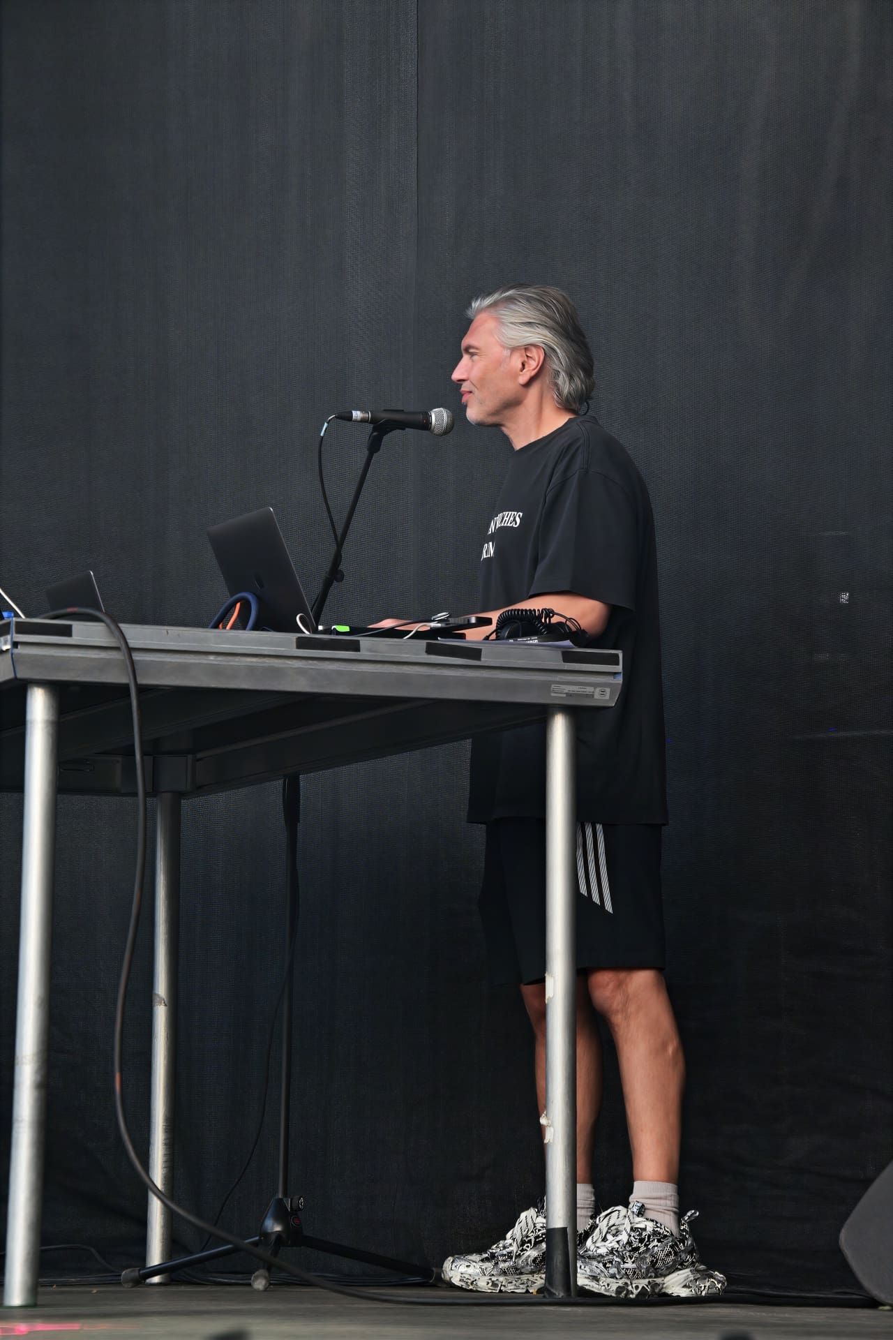 Ein Mann mit grauen Haaren steht im Profil an einem Tisch mit Laptop und Mikrofon auf der Bühne.