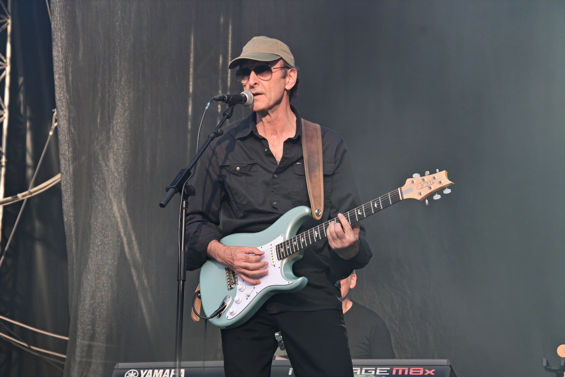 Ein Mann mit Kappe und Sonnenbrille spielt eine hellblaue E-Gitarre und singt auf der Bühne.