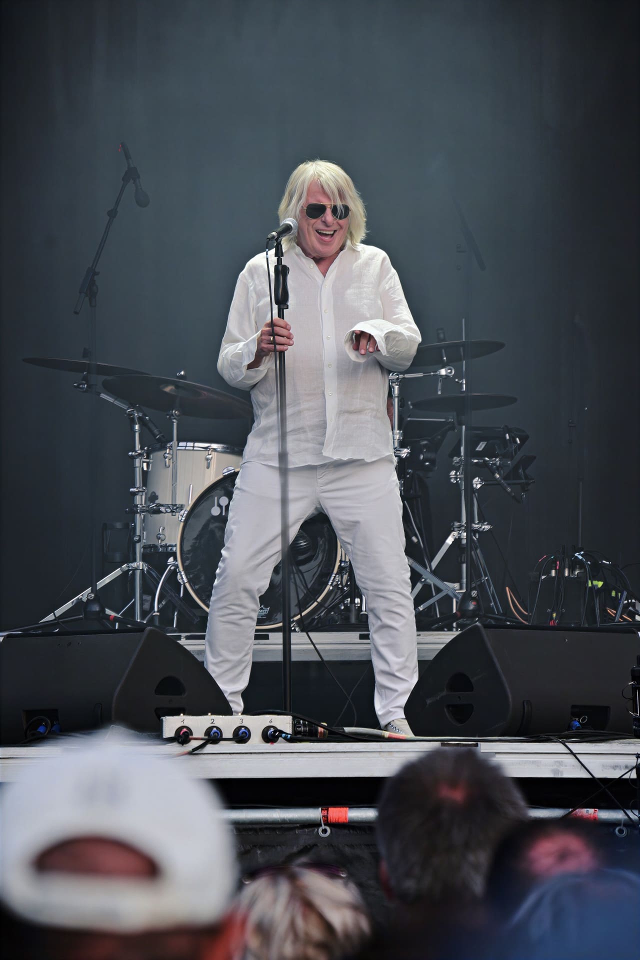 Ein Sänger mit blonden Haaren und Sonnenbrille in weißer Kleidung singt auf einer Bühne mit Schlagzeug im Hintergrund.