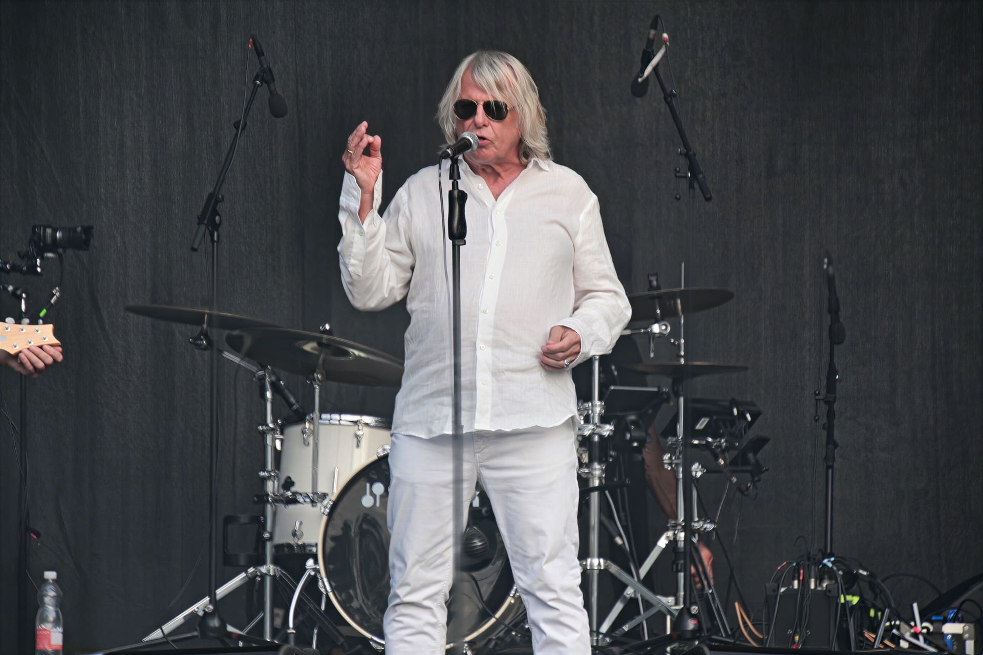 Ein älterer Sänger mit langen weißen Haaren und Sonnenbrille singt auf der Bühne vor einem Mikrofon.