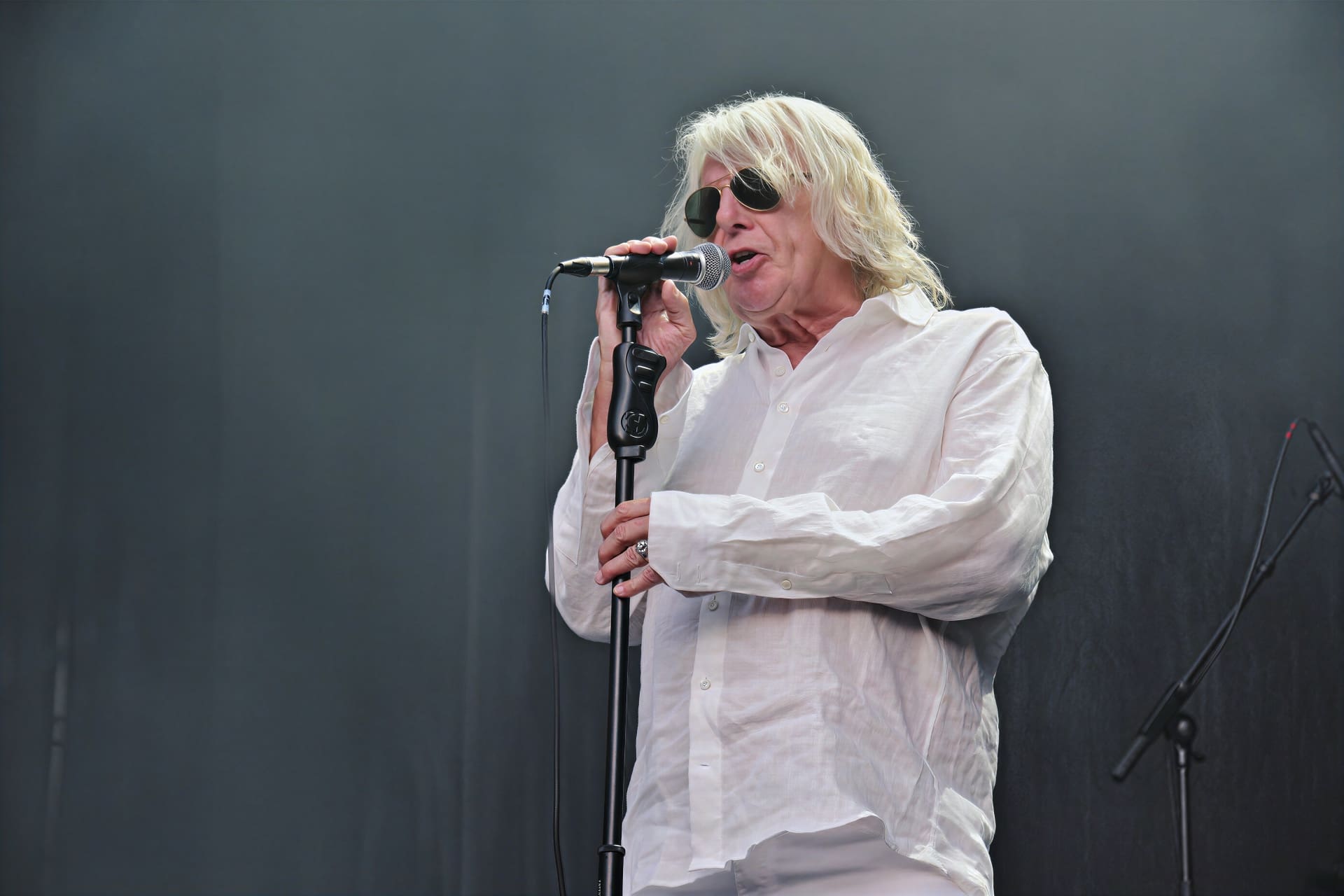 Ein Sänger mit langen weißen Haaren und Sonnenbrille singt in ein Mikrofon auf der Bühne.