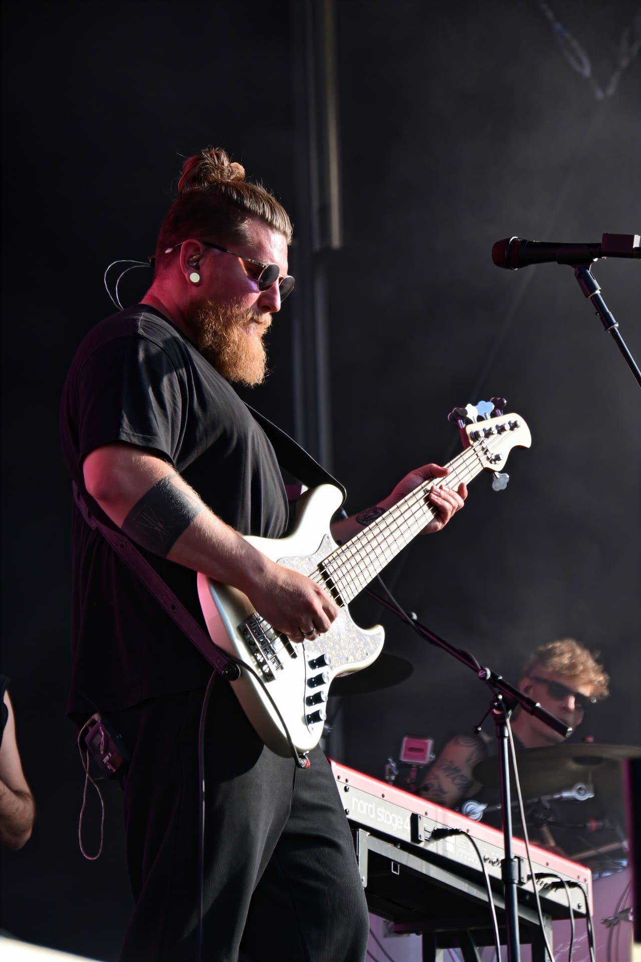 Bassist mit rotem Bart, Sonnenbrille und Dutt spielt weißen E-Bass auf der Bühne.