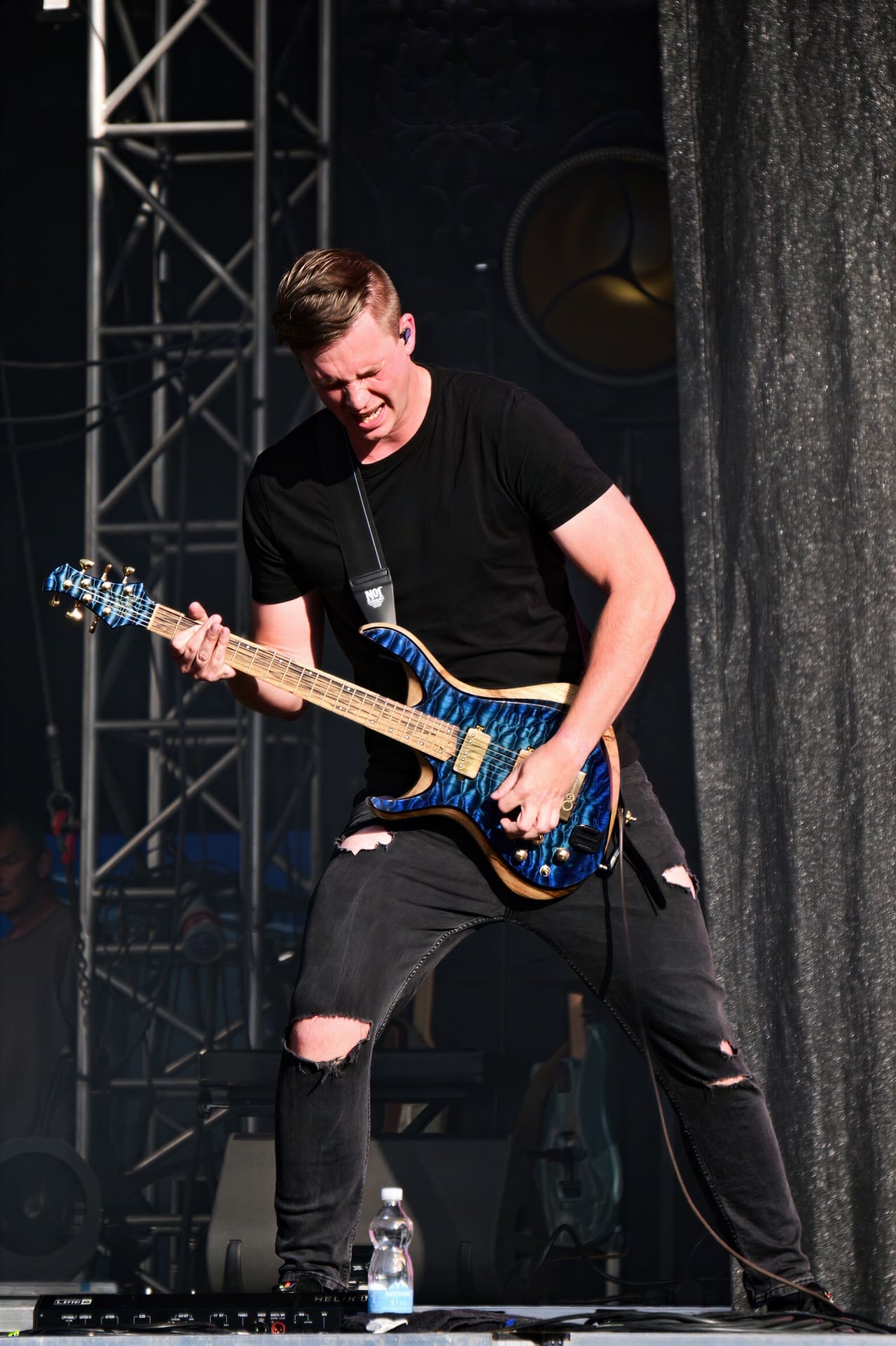 Ein Gitarrist in schwarzem T-Shirt und zerrissenen Jeans spielt eine blaue E-Gitarre auf der Bühne.