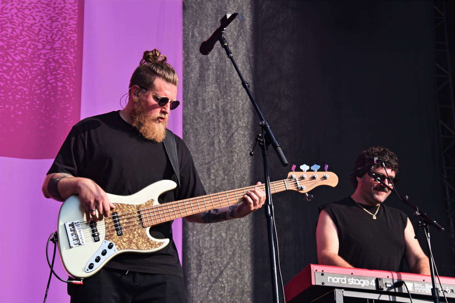 Bärtiger Bassist mit Sonnenbrille spielt auf weißem E-Bass neben Keyboarder auf der Bühne.