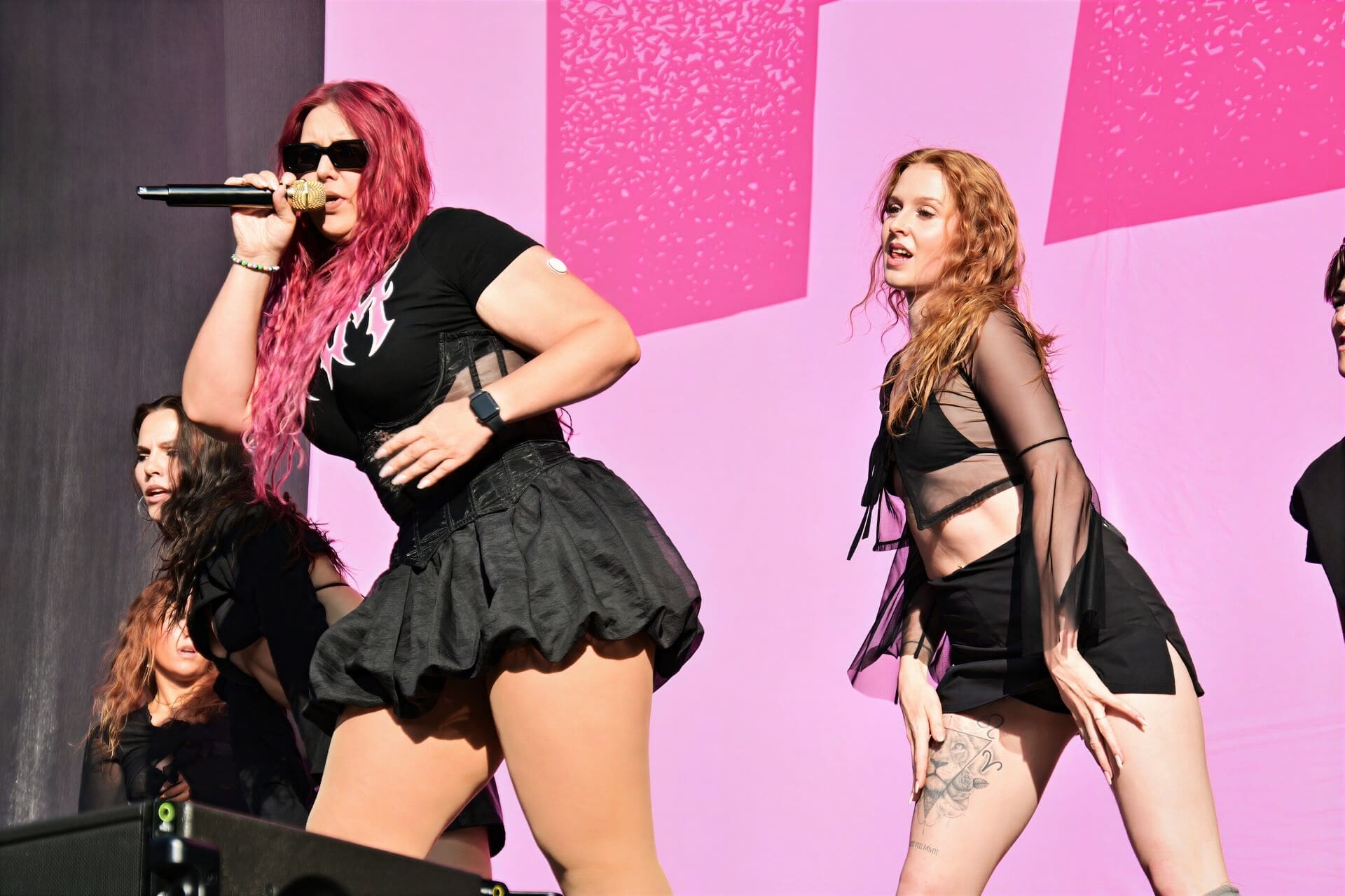 Sängerin mit leuchtend pinken Haaren und Sonnenbrille singt in ein Mikrofon auf der Bühne.