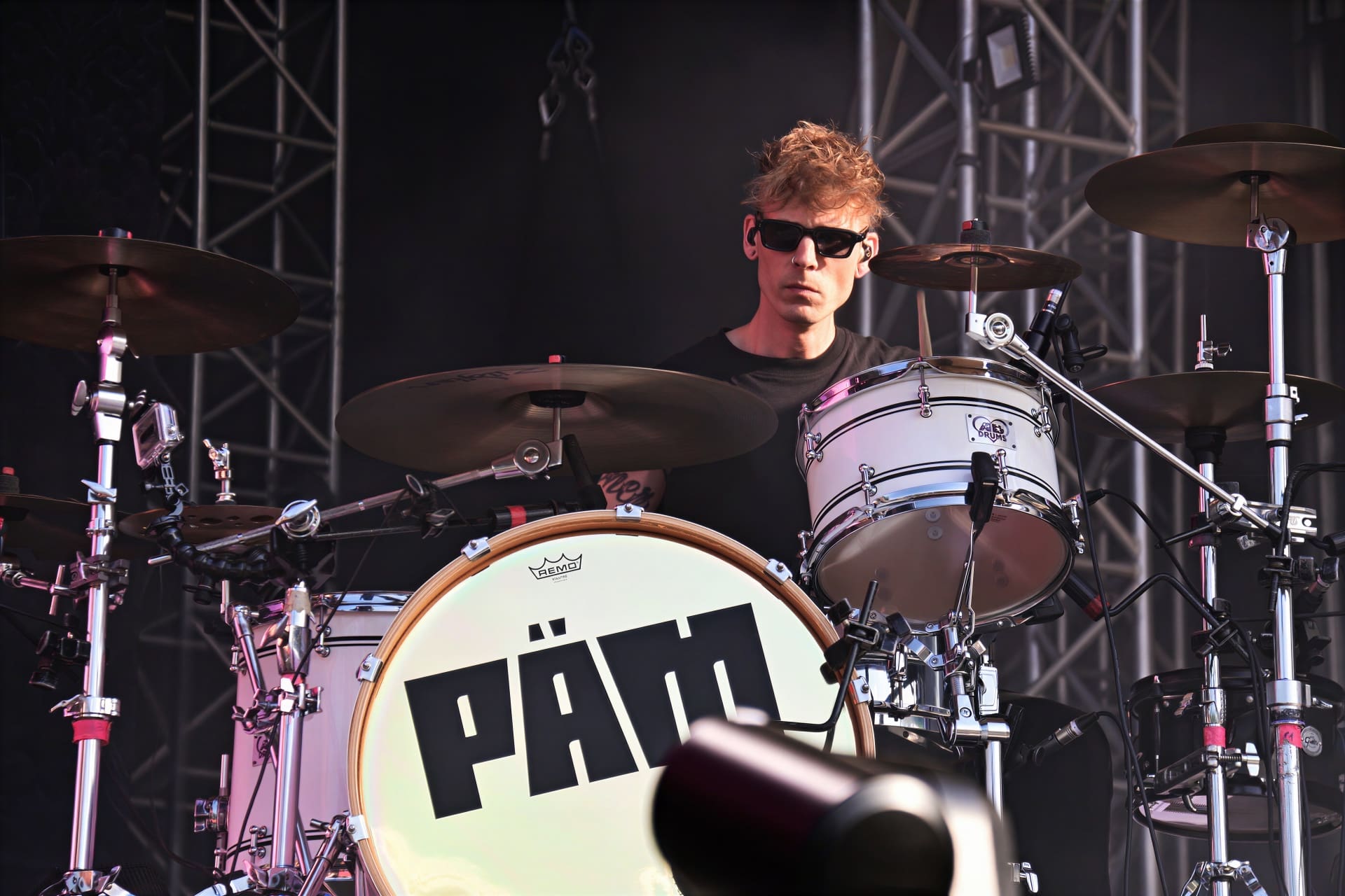 Ein Schlagzeuger mit lockigem blondem Haar und Sonnenbrille sitzt an einem Drumset mit PÄM-Aufschrift auf der Bühne.