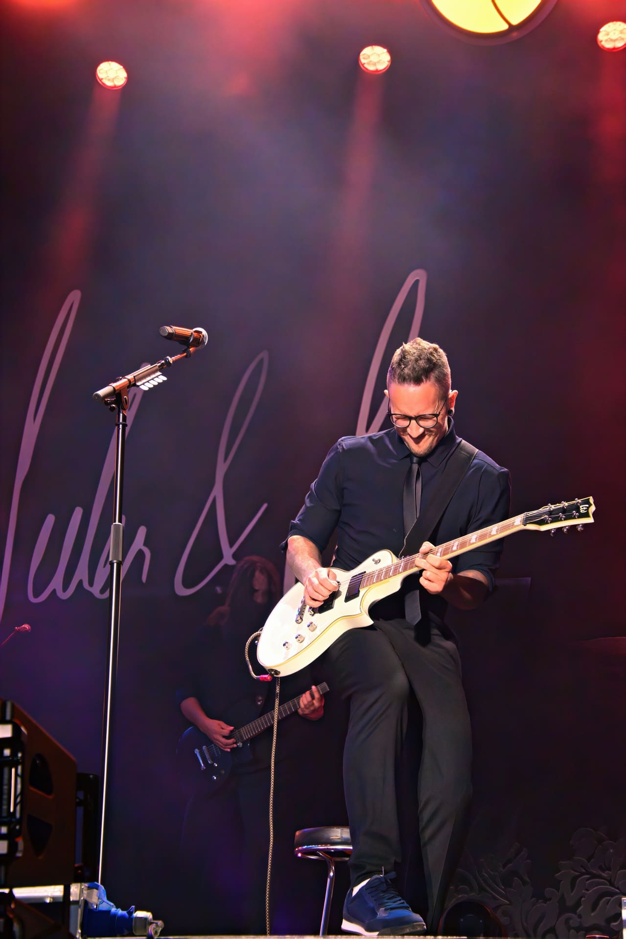 Ein Gitarrist mit Brille spielt eine weiße E-Gitarre auf einer beleuchteten Konzertbühne.