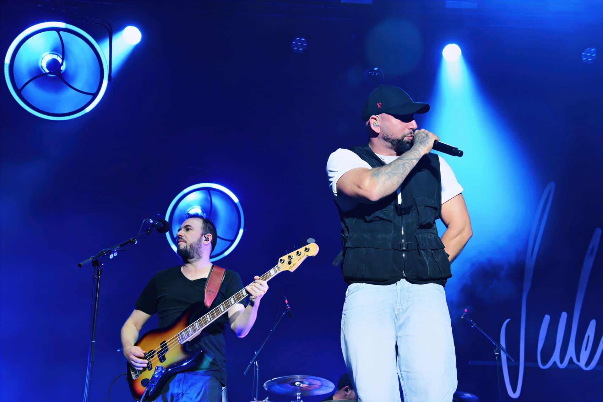 Sänger mit schwarzer Kappe und Bassist auf der Bühne bei einem Konzert unter blauer Beleuchtung