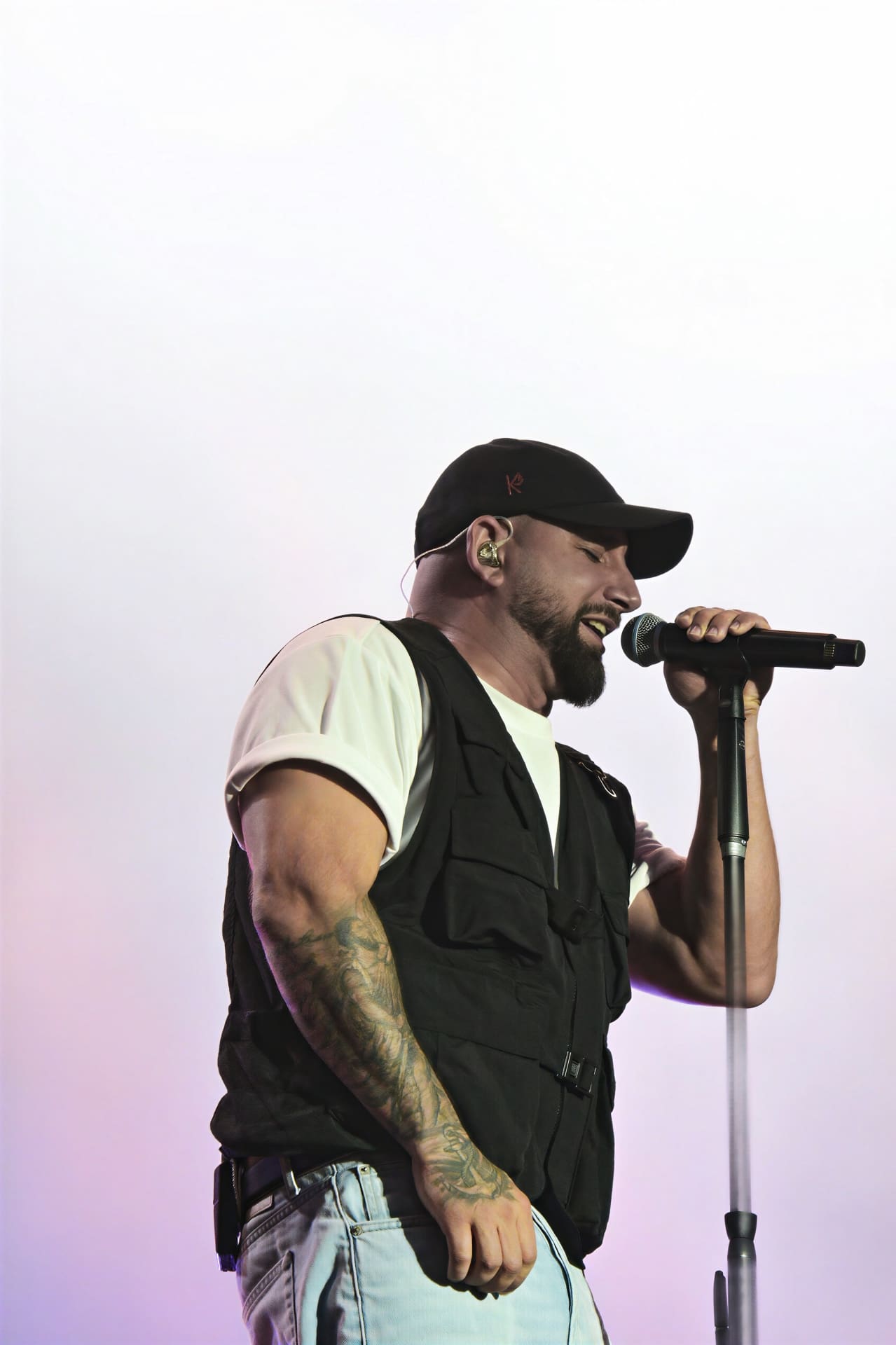 Bärtiger Sänger mit schwarzer Kappe und Tattoos am Arm singt in ein Mikrofon.