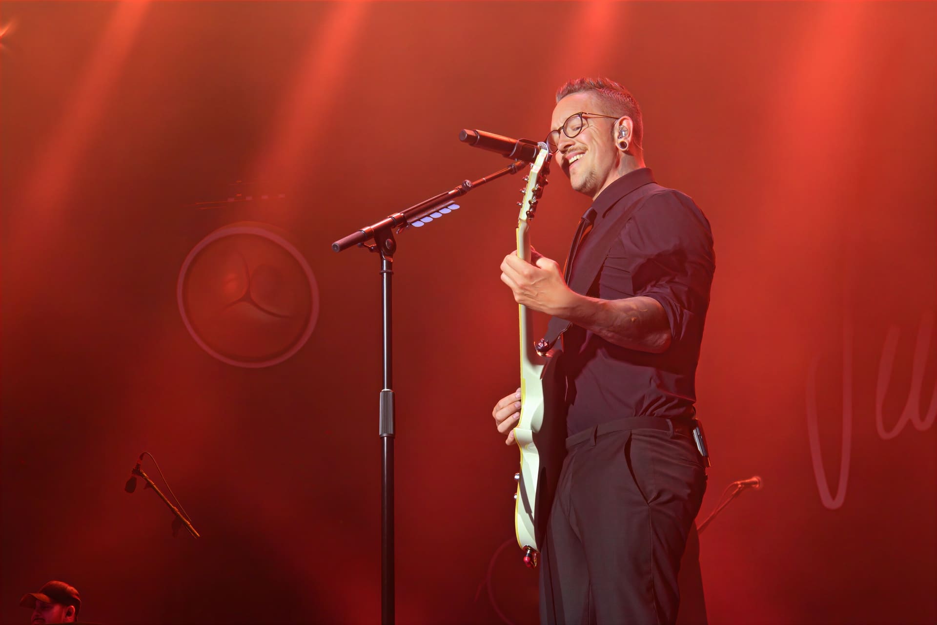 Ein Musiker mit Brille und Gitarre singt in ein Mikrofon auf rot beleuchteter Bühne.