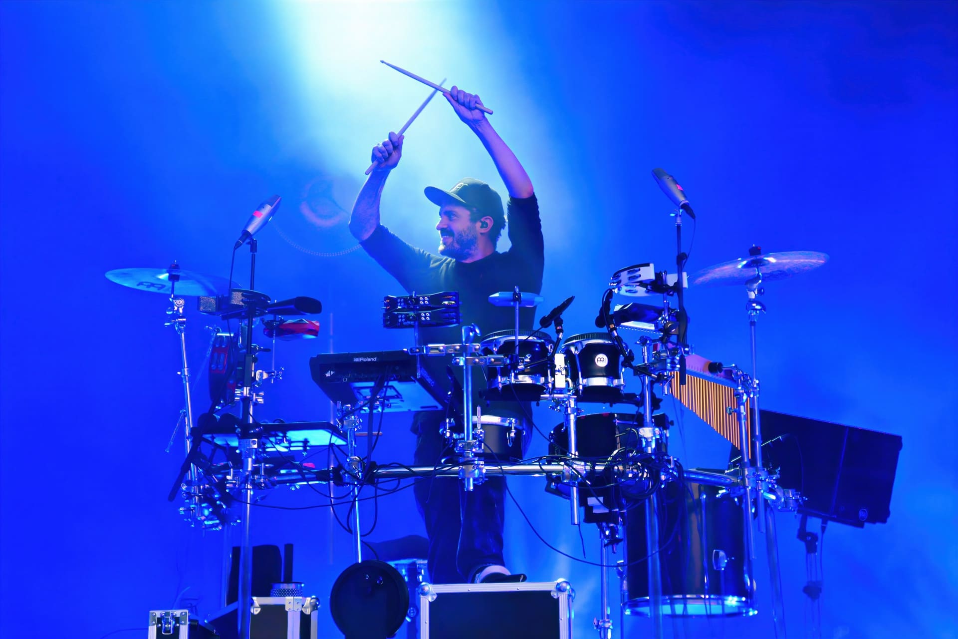 Ein bärtiger Schlagzeuger hebt die Drumsticks auf einer blau beleuchteten Bühne.
