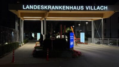 Nächtliche Ansicht des beleuchteten Eingangs zum Landeskrankenhaus Villach mit Parkplatz und Schildern