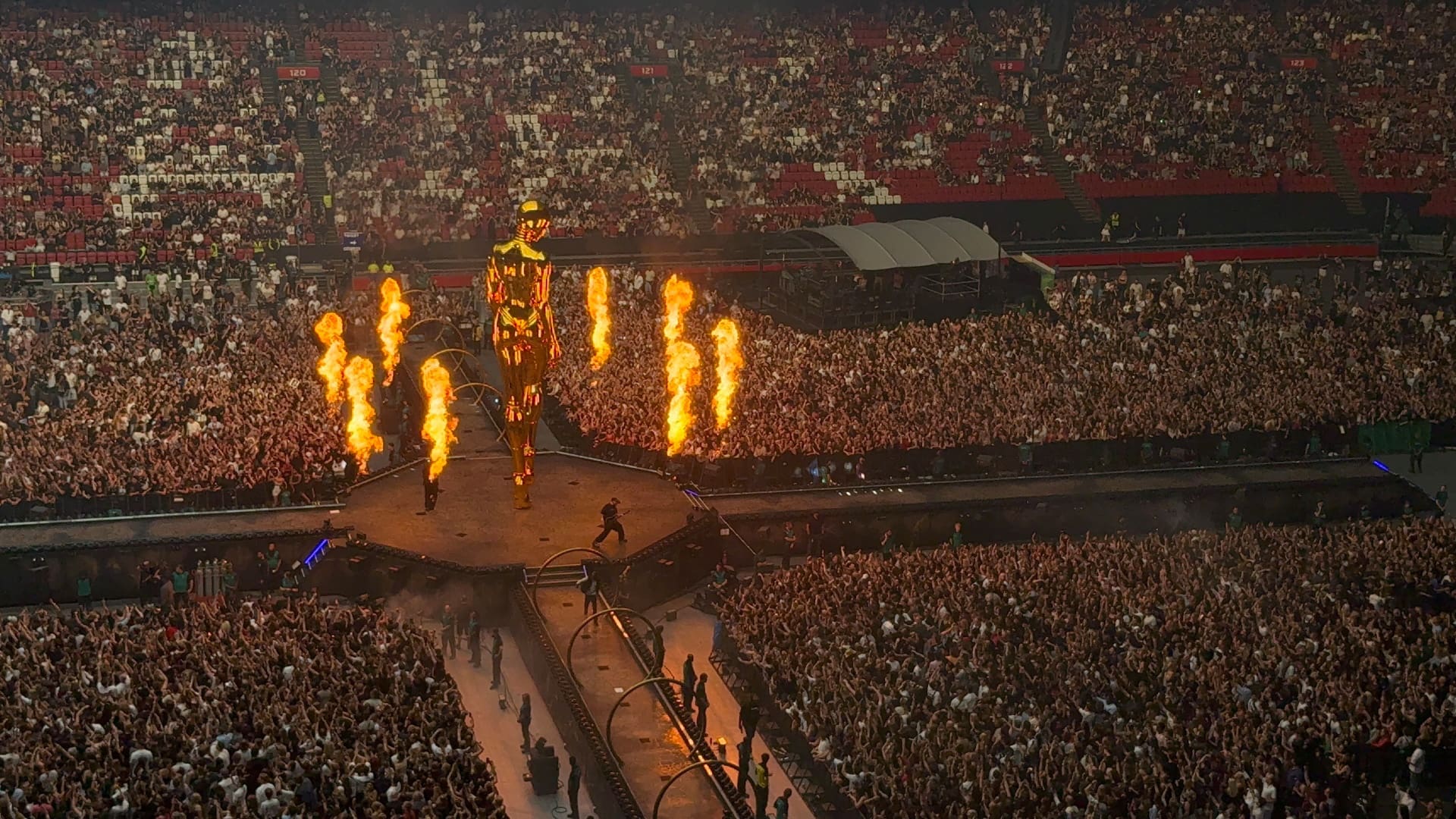 Eine große goldene Statue steht auf der Bühne eines ausverkauften Stadions mit pyrotechnischen Flammen und jubelndem Publikum.