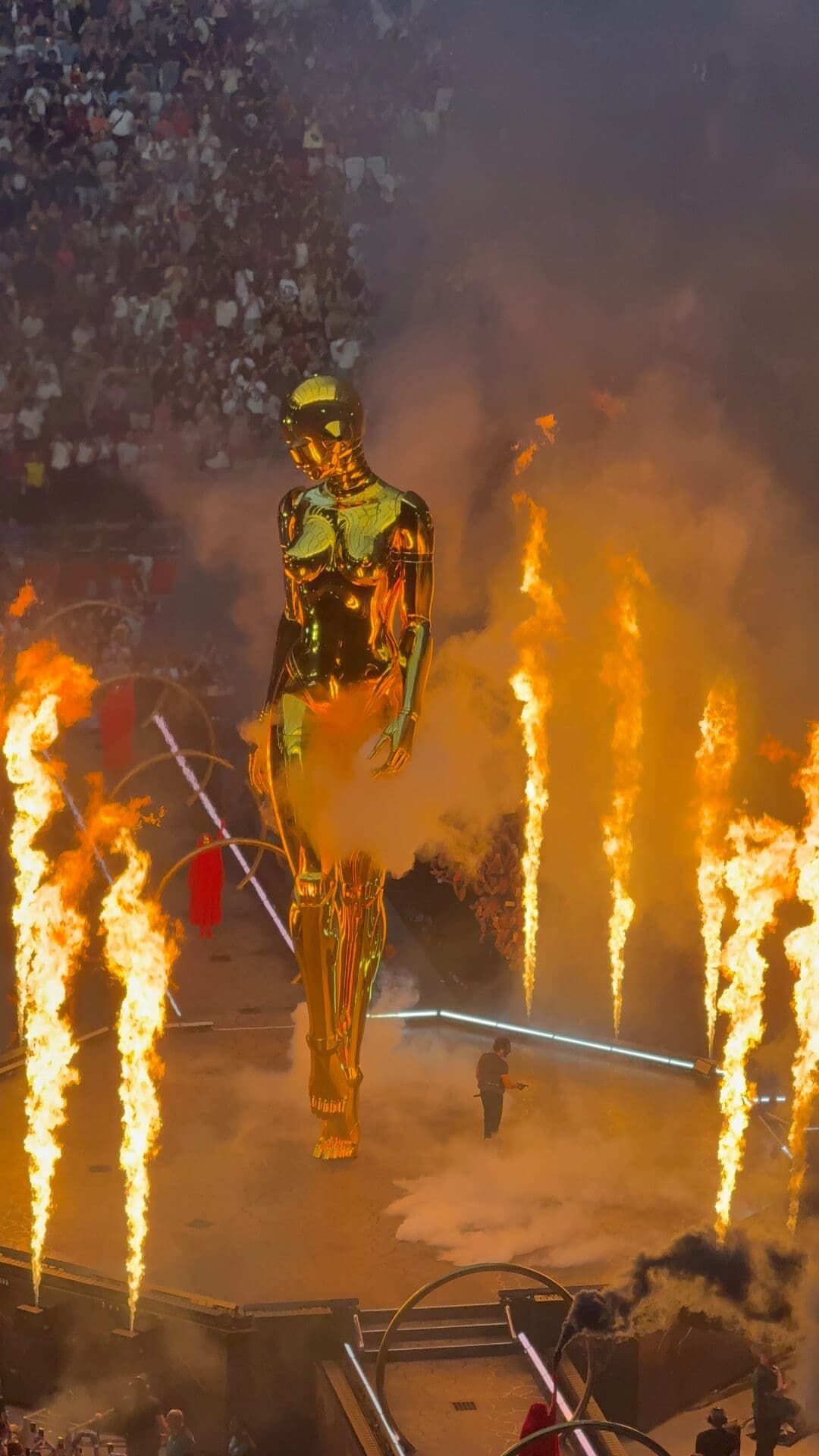 Große goldene humanoide Statue auf Konzertbühne mit Flammen Rauch und Publikum im Hintergrund