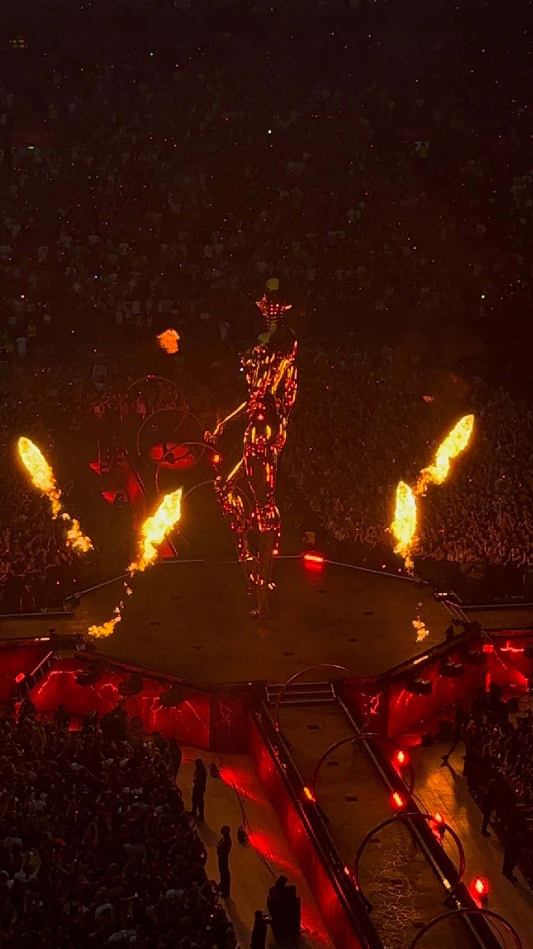 Hoch aufragende leuchtende Figur auf Konzertbühne mit Feuerstrahlen und Publikum im Stadion.