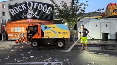Mitarbeiter in Warnkleidung reinigt mit Wasserstrahl die Straße vor dem Rock Food Stand in Villach.