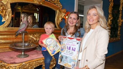 Ein Junge sitzt auf einem goldenen Tisch neben zwei Frauen mit Plakaten vor prunkvollen Spiegeln.