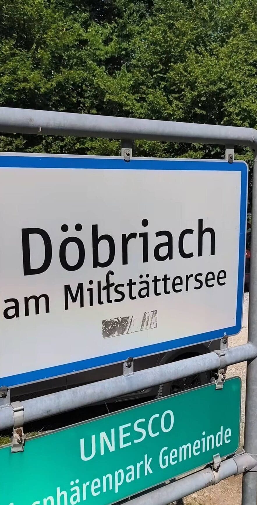 Weißes Ortsschild Döbriach am Millstättersee mit blauer Umrandung und schwarzer Beschmutzung in der Mitte