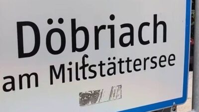 Weißes Ortsschild Döbriach am Millstätter See mit blauer Umrandung und schwarzen Verschmutzungen