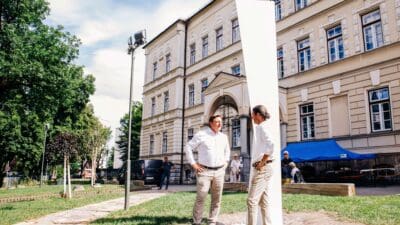 Zwei Männer in weißen Hemden stehen neben einer hohen weißen Stahlskulptur im Park vor einem Gebäude.