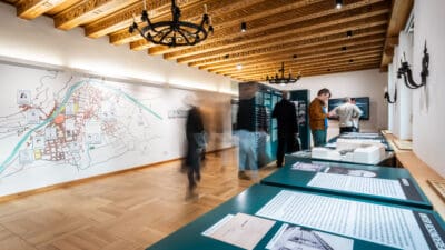 Besucher in einem Ausstellungsraum mit großer historischer Stadtkarte an der Wand und Modellen auf Tischen.