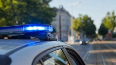 Nahaufnahme eines leuchtenden blauen Blaulichts auf dem Dach eines silbernen Polizeifahrzeugs in einer Straße mit Gleisen.