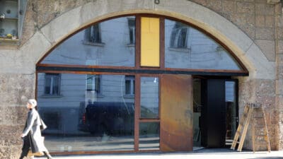 Eine Frau in grauem Mantel und Mütze geht an einem großen Rundbogen mit Glas und Leitern vorbei.